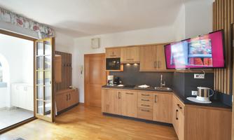 Modern kitchen with wooden cabinets and a refrigerator. A television is mounted on the wall, and large windows provide plenty of natural light.