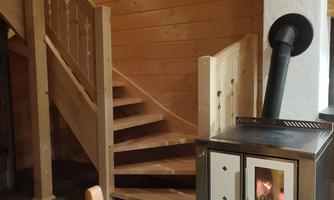 A cozy room with a wooden staircase and a modern stove. The floor is covered with dark tiles, and the walls are made of wood.