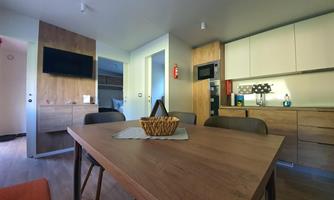 A modern living kitchen with a wooden table and chairs. In the background, a television and a well-equipped kitchen can be seen.