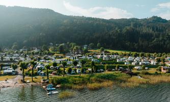 A campsite by the water with numerous motorhomes. In the background, green hills and forests stretch out.
