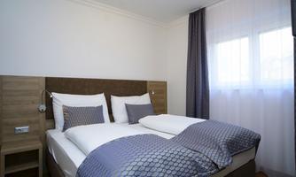 A modern bedroom with a double bed and light curtains. The walls are kept neutral, and a bedside table is located next to the bed.