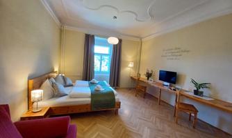 A bright, elegant room with a double bed and a comfortable armchair. The room has large windows, a small desk, and a modern TV unit.
