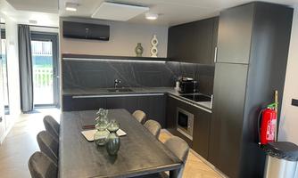 A modern kitchen with a dining table and elegant chairs. The kitchen furnishings are contemporary with a black wall and large windows.