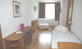 A simple room with a single bed, a desk, and a chair. A window lets in daylight and provides a bright atmosphere.