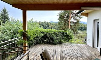 A wooden terrace with a cozy chair. The view shows lush foliage and hills in the background.
