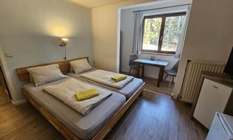A simple room with two single beds, a small seating area, and a window. The room has a bright, friendly atmosphere with minimalist design.