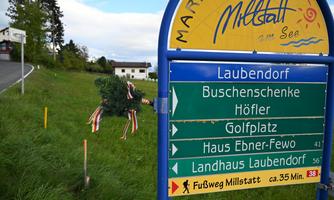 A signpost with various directions to places in the vicinity, including the leaf village and golf course. In the background, green meadows and a residential building are visible.