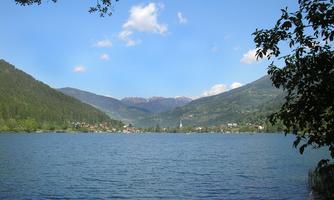 A clear lake surrounded by green mountains and forests. The sky is blue with some white clouds.