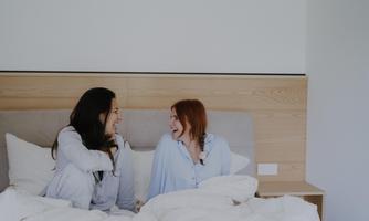 Two women are laughing in bed and enjoying a relaxed moment. They are wearing cozy pajamas and surrounded by pillows and blankets.