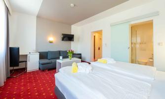 A spacious hotel room with two beds, a table, and a cozy sofa. The modern furnishings and bright light create an inviting atmosphere.