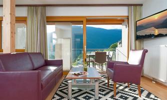 A modern living room with a dark purple sofa and an armchair. The large window front offers a view of the mountains and the terrace.