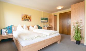 A cozy bedroom with a large bed and bright walls. There is a wardrobe, a nightstand, and a beautiful plant in the corner.