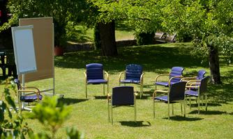 A circle of blue chairs stands on a green meadow. In the background, a flip chart and trees can be seen.