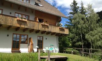 A cozy wooden house with a balcony and flower boxes. The garden is green and surrounded by trees.
