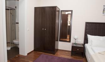 A simple hotel room with a bed, a nightstand, and a wardrobe. In the background, a bathroom and a mirror are visible.