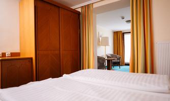 A cozy hotel room with a large bed and a wardrobe. Through the open door, you can see another room with a seating area and windows.