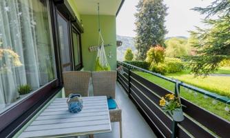 A cozy balcony with two chairs and a table. In the background, a garden with trees and flowers can be seen.