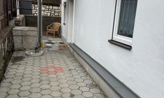 A paved path next to a house with a terrace and a chair. The wall is white and the sky is cloudy.