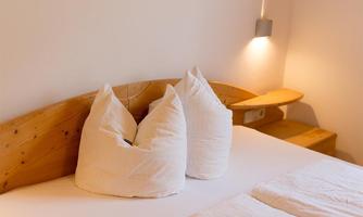 A cozy bedroom with a wooden bed and two large pillows. The wall is painted in soft colors, and a small lamp provides warm light.