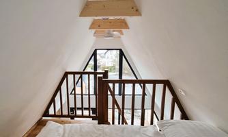 A cozy bedroom in the attic with a wooden railing and a large window front. The walls are simple and bright, creating an inviting impression.