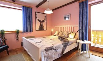 A cozy bedroom with a white bed and a large window. The walls are painted in shades of pink and there are decorative curtains and plants.