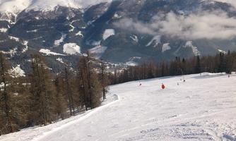 A snow-covered mountain landscape with heavy snowfall and trees in the foreground. In the background, majestic mountains and a clear sky can be seen.