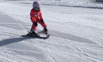 A small child is skiing on a groomed slope. The sky is clear and the sun is shining.