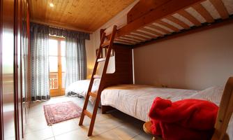 A cozy bedroom with a bunk bed and a single bed. The room has windows with curtains and wood paneling on the ceiling.