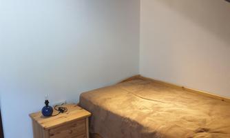 A simple bedroom with a bed and a wooden nightstand. The walls are painted light, and there is a small picture on the wall.