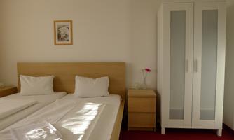 A simple and bright bedroom with two beds and a chest of drawers. A white wardrobe is against the wall, and a picture hangs above the bed.
