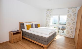 A modern bedroom with a double bed and colorful pillows. Large windows provide plenty of daylight and a view outside.