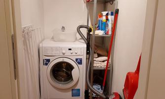 A small utility room with a washing machine, a vacuum cleaner, and various cleaning supplies on a shelf. A drying rack is against the wall.