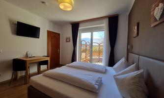 A modern room with a double bed and a large window leading to a balcony with a view of the outdoors. Equipped with a table, a television, and stylish decoration.