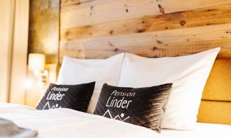 A cozy bed with white pillows and a wooden paneling in the background. The pillows are adorned with the inscription "Pension Linder."