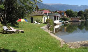 A picturesque lakeside landscape with green grass, deck chairs, and a colorful parasol. In the background, cozy houses and a pier can be seen.