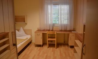 A simple room with a bed, a desk, and a window. The room has light walls and a wooden floor.
