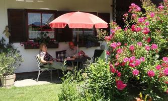 A cozy outdoor area with a table and two people sitting under a red umbrella. Pink roses are blooming all around and the grass is green.