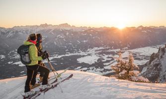 Two skiers stand on a snow-covered slope and look at the sunset over the mountains. As the sun sets, the sky glows in warm colors.
