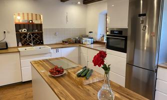 A modern kitchen with a wooden worktop, fresh produce, and a vase with red flowers. In the background, a refrigerator and a small bar are visible.