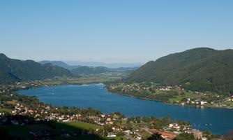 A picturesque landscape with a tranquil lake, surrounded by hills and small villages. The sky is clear and blue, perfect for a relaxing day in nature.