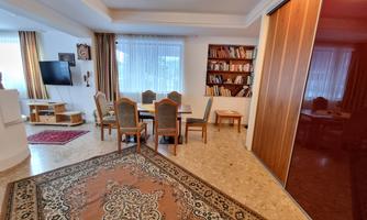 A bright living room with a large table and chairs. In the background, shelves with books and a television are visible.