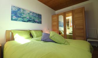 A cozy bedroom with a large bed and green bedding. A painting of water lilies hangs on the wall, and in the background, there is a wardrobe.