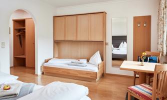 A simple room with wooden shelves, a bed, and a table. The walls are light and there is a large mirror.