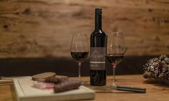 A bottle of red wine is on a table, flanked by two wine glasses. Next to the bottle are a wooden board and some delicacies.