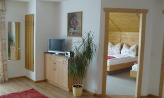 A cozy room with a television, a wardrobe, and a large mirror. In the background, you can see a bedroom with a wooden bed and decorative cushions.