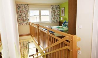 A bright hallway with wooden railings and a view of a cozy bedroom. The walls are painted in friendly colors, and the windows are decorated with colorful curtains.
