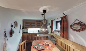 A cozy kitchen with wooden shelves and a dining table. Decorative elements hang on the walls and the windows are adorned with curtains.