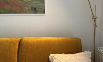 A modern living room with a yellow sofa and a white pillow. A painting of a landscape with a blue sky and green fields hangs on the wall.