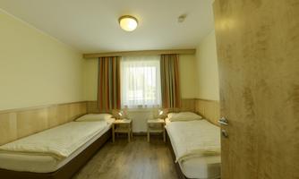 A simple guest room with two single beds and a window through which daylight enters. The walls are light and the furniture is made of wood.