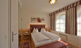 A bright and cozy bedroom with two single beds and a red seating area. Large windows let in plenty of daylight.